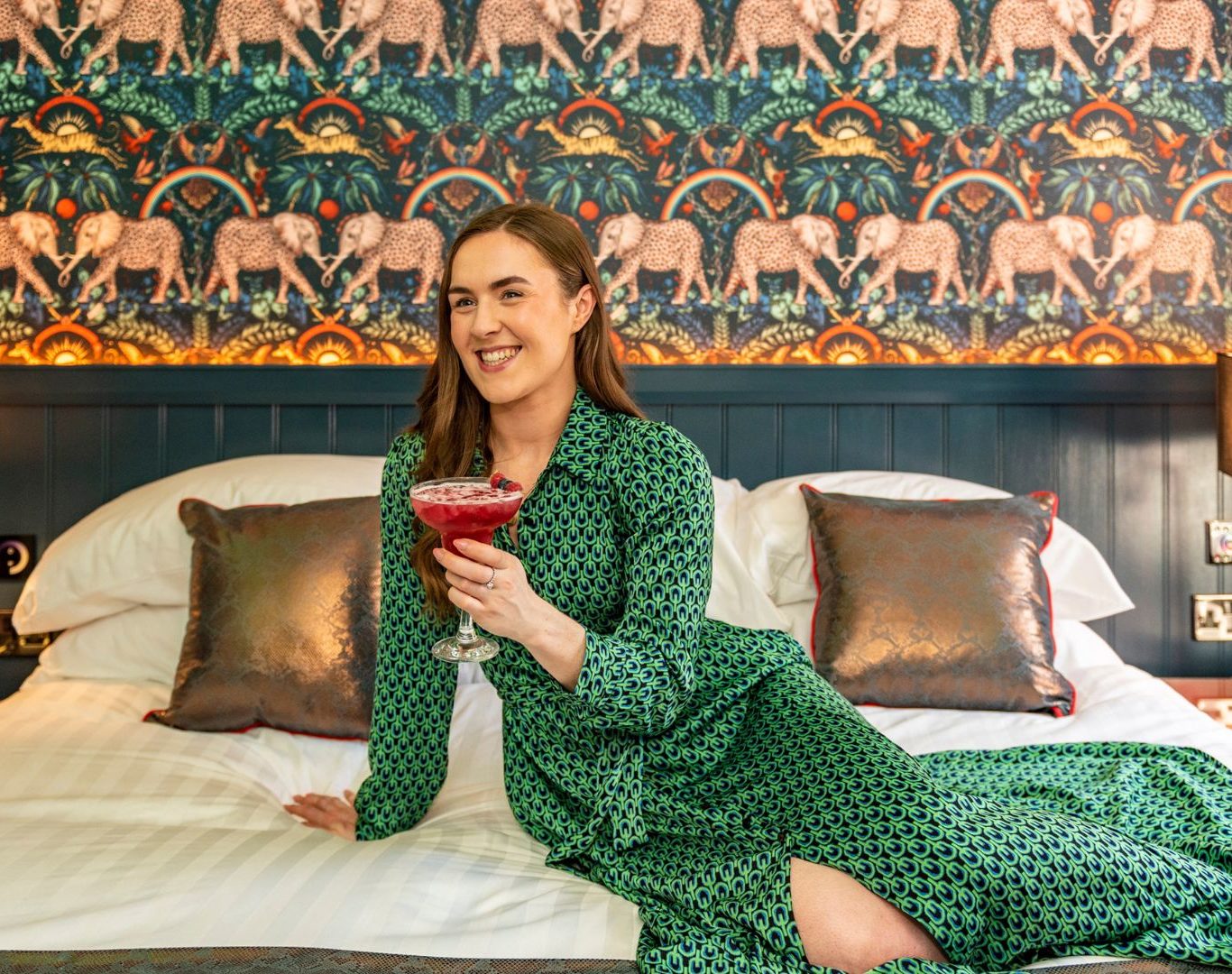 A woman in a green dress lying on a bed in Crown Quarter Hotel holding a red cocktail. Book direct for the best special offers for hotels in Ireland